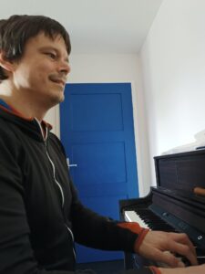 Mann spielt Klavier. Musiker mit dunkler Jacke und Klaviertasten. Blaue Tür im Hintergrund.