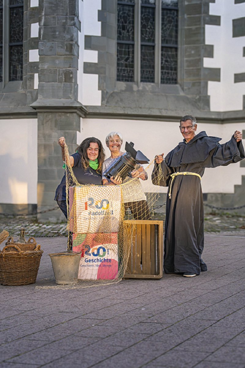 Drei Personen posieren vor der Kirche St. Radolt. Kisten, Netz, Mönchskutte, Lampe. Radolfzell Geschichte.