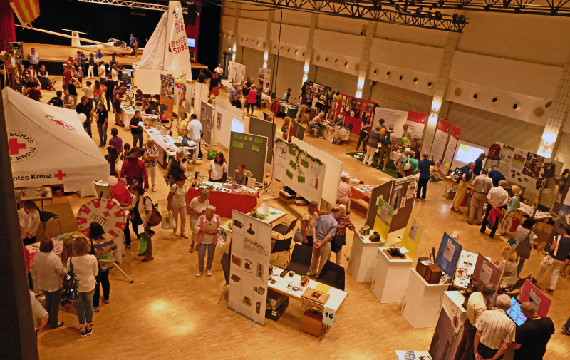 event-a01e27fb-52ad-4ad5-9594-fa69d4ad0ba1-1 Messe-Überblick: Menschen an Ständen, Rotes Kreuz Zelt, Segelboot auf Bühne. Messeveranstaltung mit Publikum.