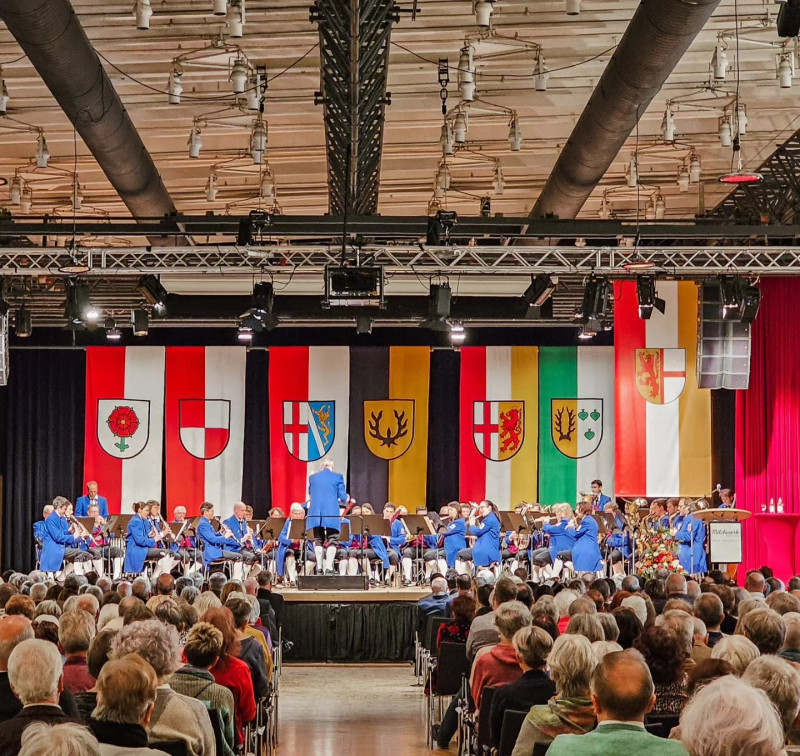 Konzert einer Musikkapelle vor Publikum. Bühne mit Wappen, Dirigent und Musiker in blau.