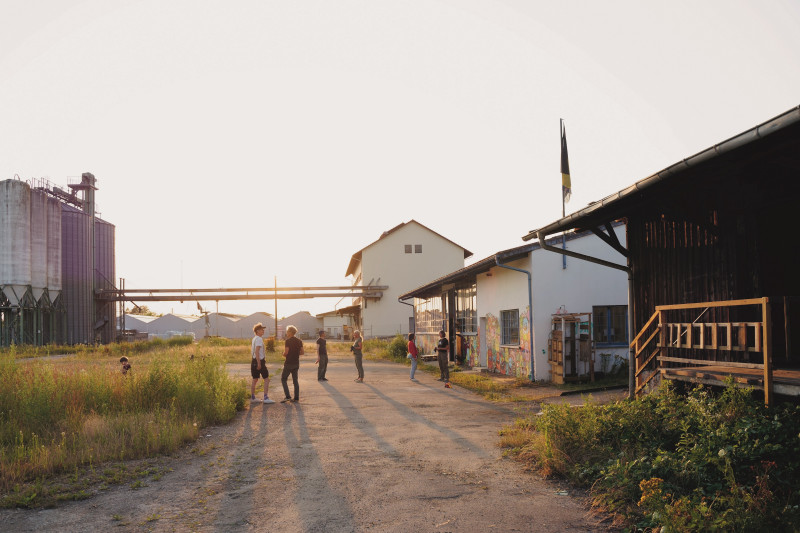 Gruppe von Menschen vor Industriegebäude mit Getreidesilos und Lagerhallen im Abendlicht.
