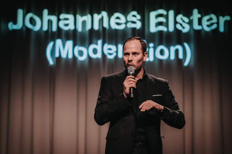 Johannes Elster, Moderator im schwarzen Anzug, spricht ins Mikrofon auf der Bühne.