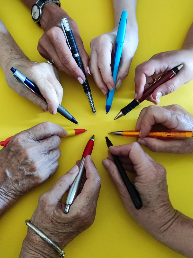 Mehrere Hände halten Stifte verschiedener Farben auf gelbem Grund, die auf einen Punkt in der Mitte zeigen.