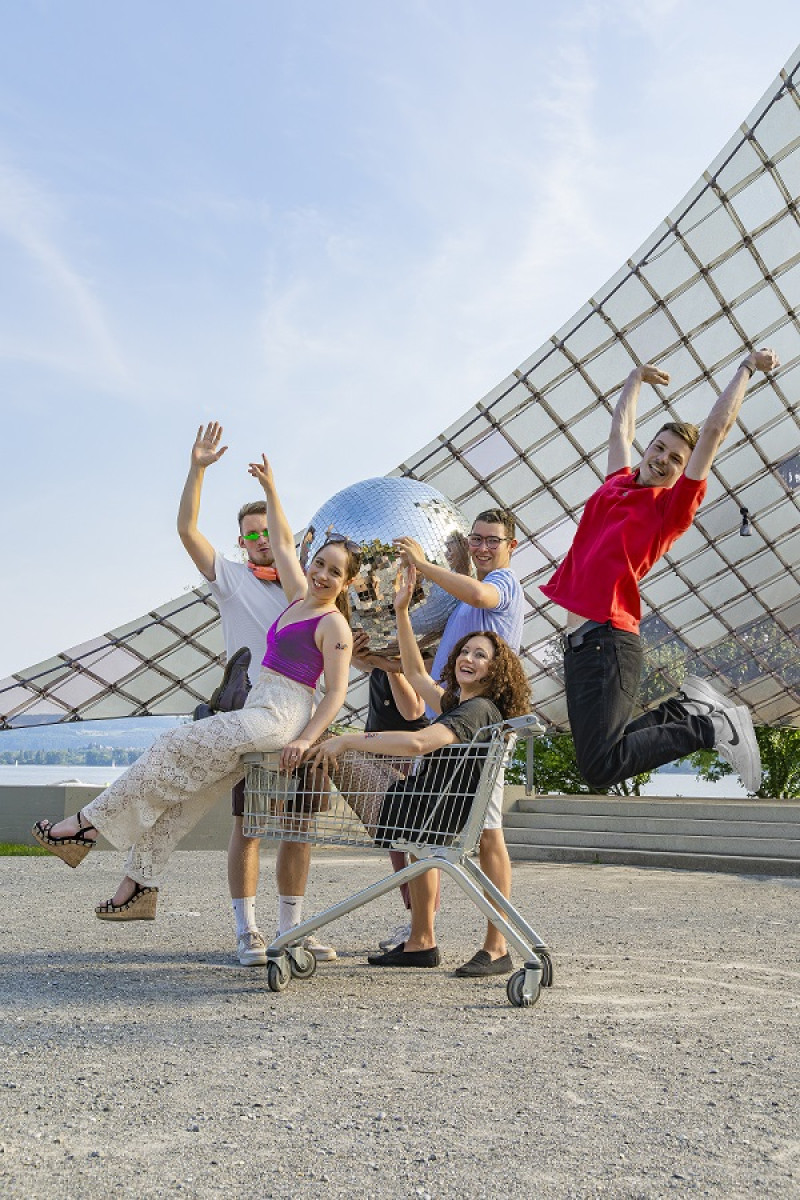 Five friends having fun with a shopping cart and disco ball in front of a glass building.
