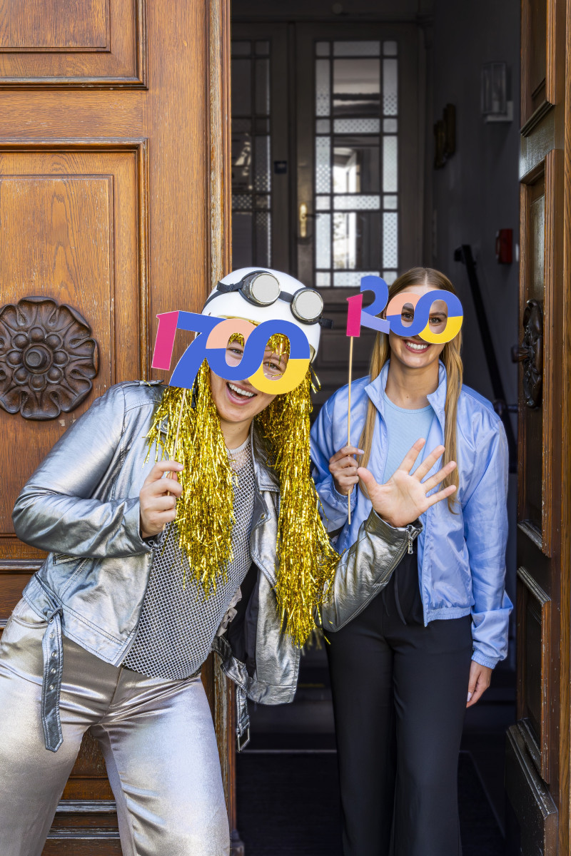 Zwei feiernde Frauen mit 700-Jahre-Jubiläumsbrillen stehen in einer Tür. Party, Spaß und goldene Perücke.