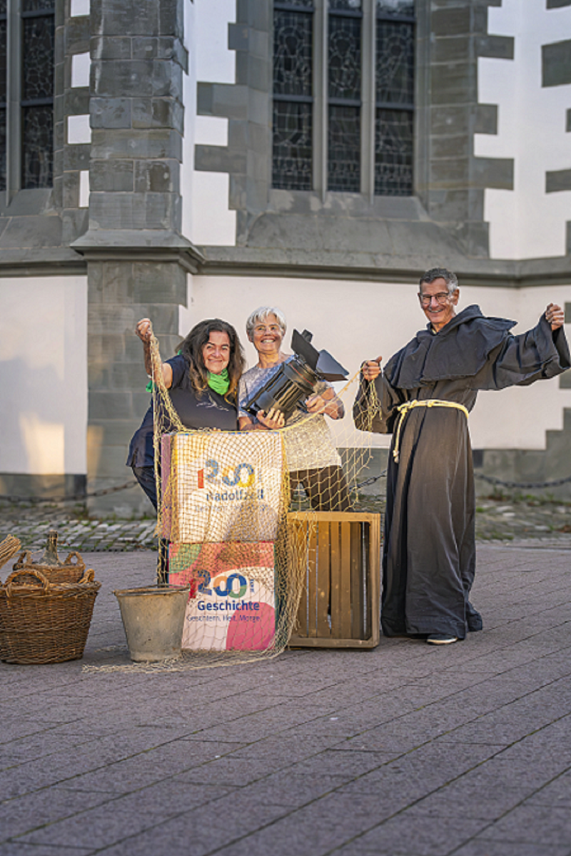 Drei Personen inszenieren 1200 Jahre Radolfzell mit Requisite vor historischer Kirche. Mönch, Scheinwerfer, Fischernetz.
