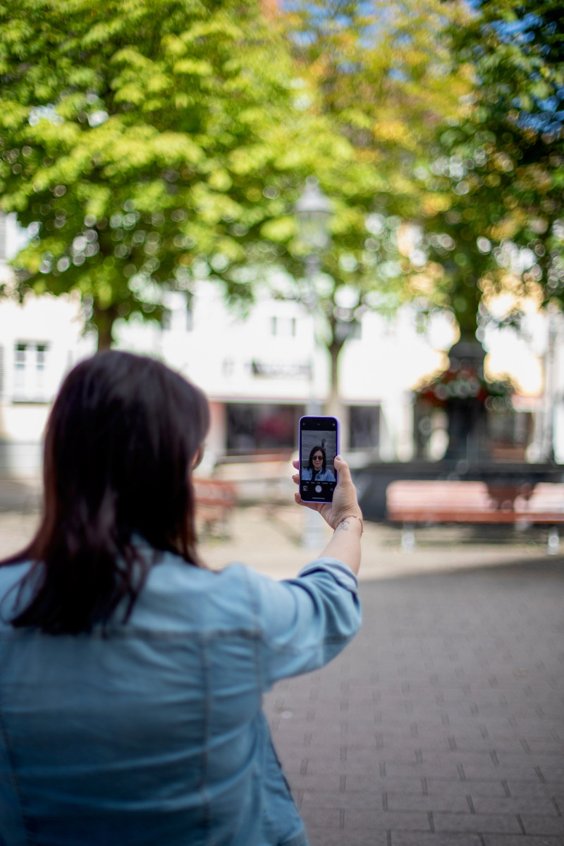 Frau macht Selfie mit Smartphone in der Stadt. Bäume und Gebäude im Hintergrund. Reisefotografie in Deutschland.
