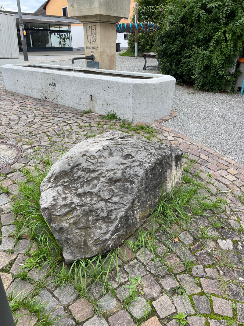 Markelfingen Brunnen mit historischem Stein und Inschrift, umgeben von Kopfsteinpflaster und Grün.